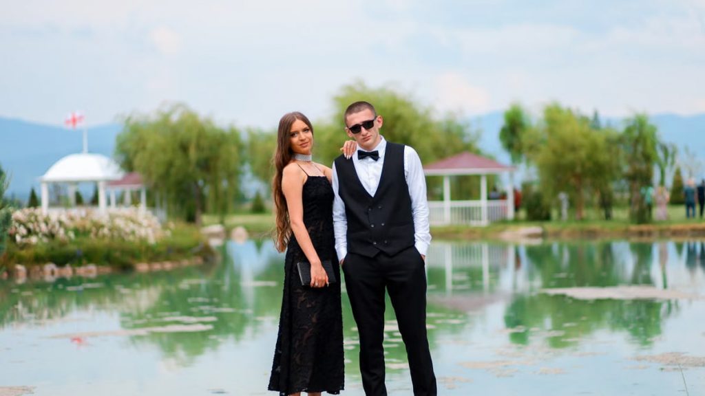 A couple in formal attire poses elegantly by a serene lake, capturing a romantic moment.